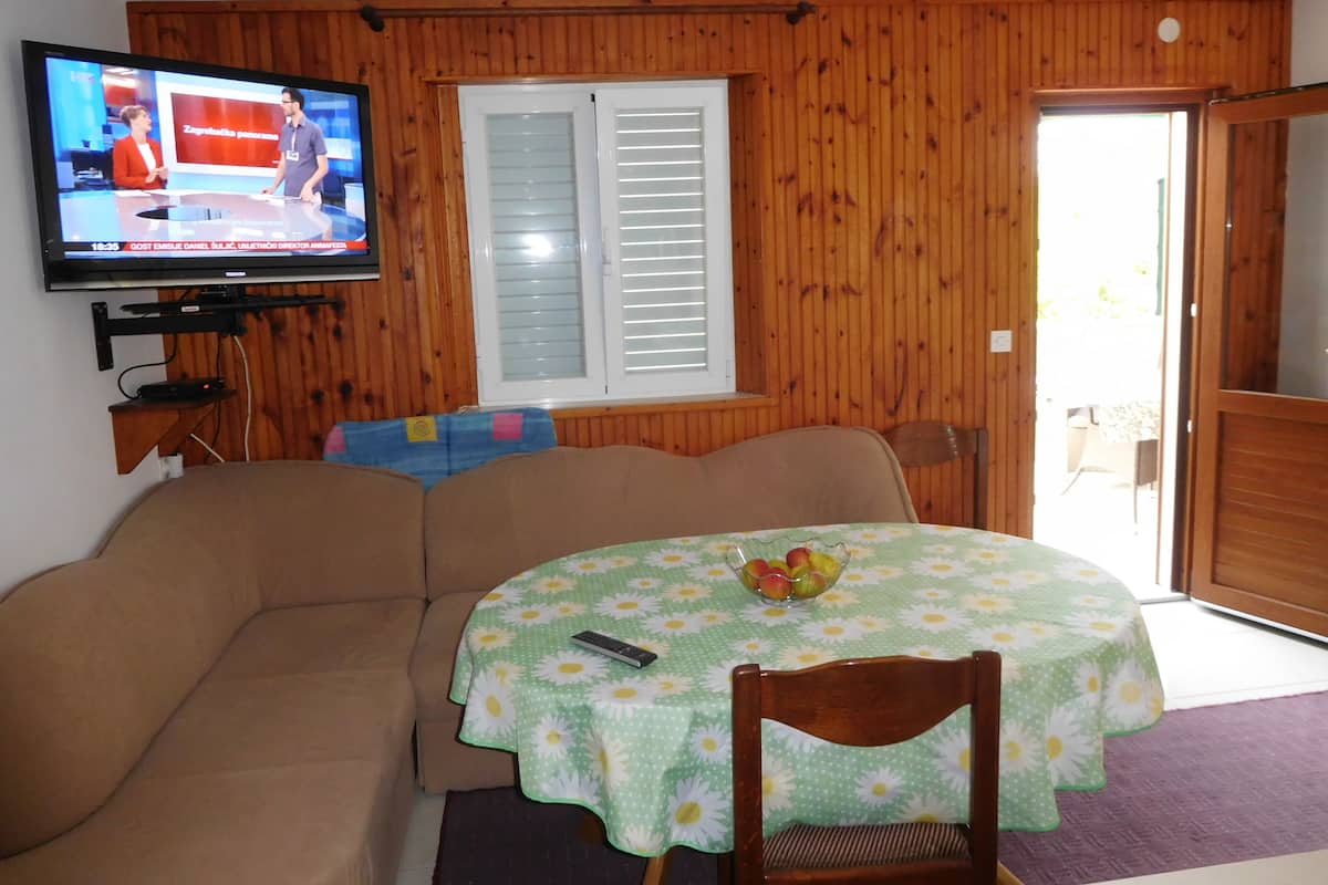 A living room with a wooden paneled wall, a television mounted above a sofa, and a dining area with a table covered in a floral tablecloth.