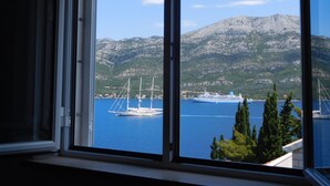 A view through a window of a coastal area with a large cruise ship docked in the water, mountains in the background, and a few sailboats. - Apartments Ivancevic (Korcula)