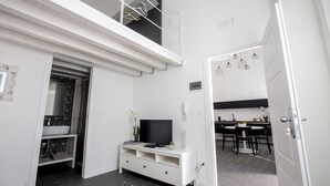 Un interno moderno e luminoso, con una TV su un mobile bianco, un'area pranzo con tavolo e sedie, e una scala che conduce a un piano superiore.