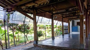A covered outdoor area with a brick floor, wooden beams, and a view of lush greenery through large windows. - Griya Jagadhaya (Sewon)