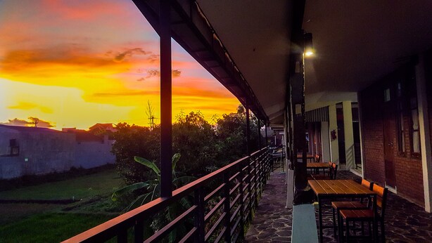A balcony with a view of a sunset, outdoor seating, and a building in the background. - Griya Jagadhaya (Sewon)