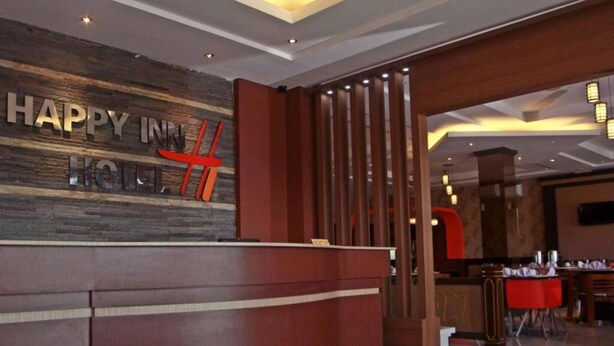 A reception area with a wooden reception desk, a 'Happy Inn' sign, and a glimpse of a dining area with tables and chairs. - Happy Inn Hotel (Kendari)