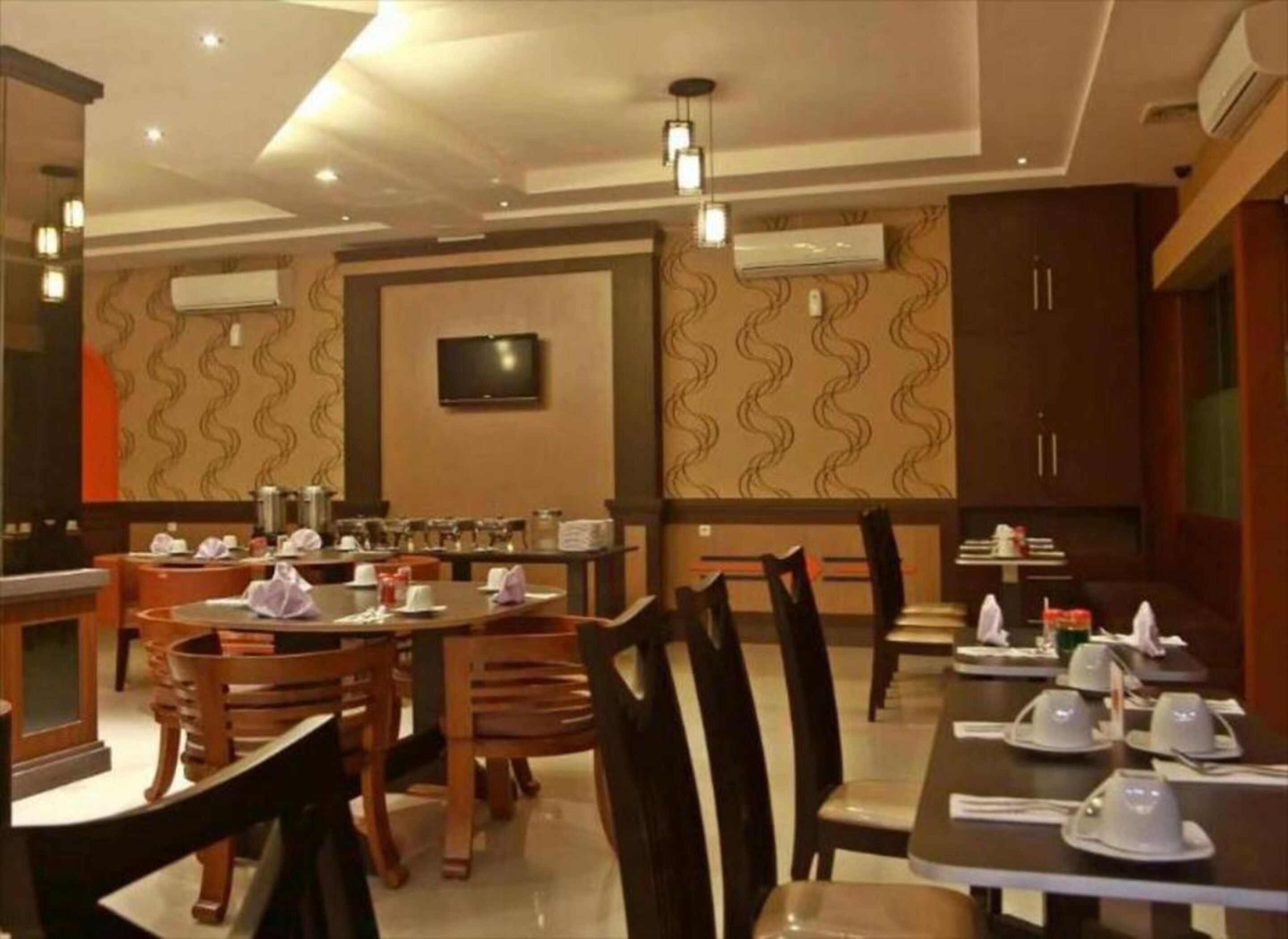 Um restaurante com várias mesas e cadeiras, uma televisão fixada na parede e unidades de ar condicionado no teto.