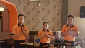 Three staff members in orange uniforms standing in a dining area with a patterned wall and an air conditioning unit above.