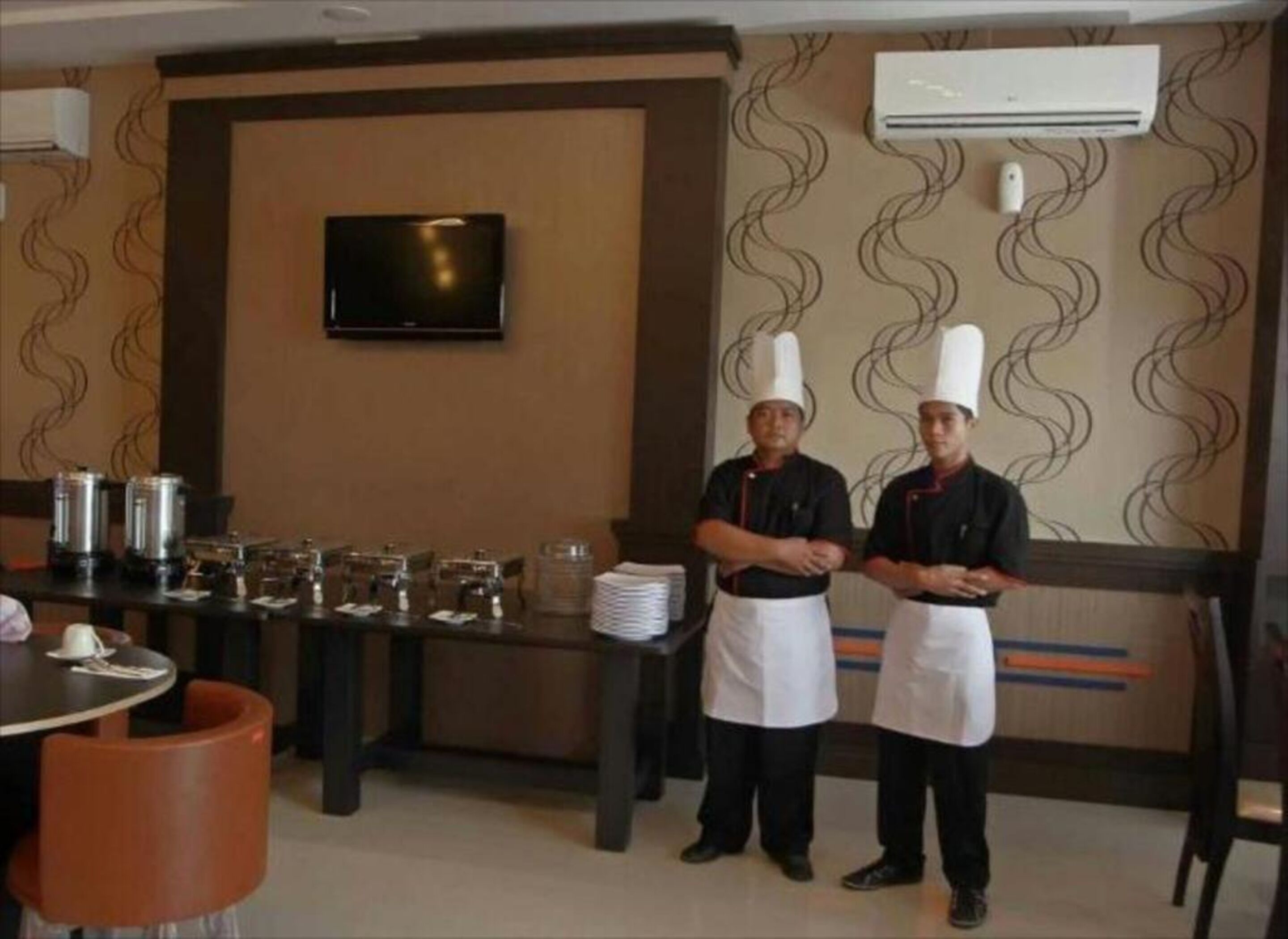 A restaurant interior with chefs standing by a buffet table, a television on the wall, and an air conditioning unit.