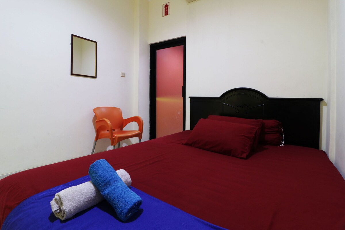Ein Einzelbett mit roter Bettwäsche, ein blau-weißes Handtuch und ein rotes Kopfkissen. Es gibt ein schwarzes Kopfteil, eine rote Tür und einen kleinen orangefarbenen Stuhl.
