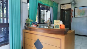 A reception desk with a wooden finish and a black diamond-shaped design. There is a window with green curtains, a map on the wall, and a fan near the door. - Wisma Flamboyan (Surabaya)