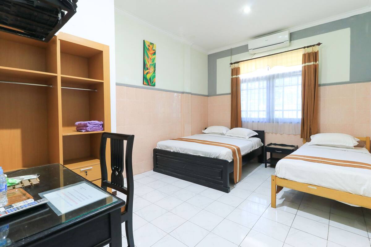 A hotel room with two beds, a desk with a chair, a wardrobe, and a bathroom with a window and curtain.