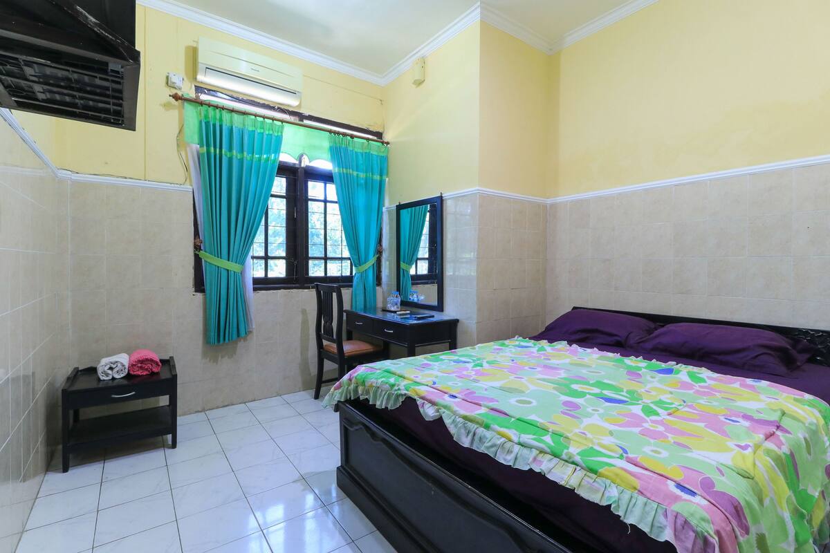 A bedroom with a bed, a desk, a chair, a small table with towels, a window with curtains, and an air conditioning unit.