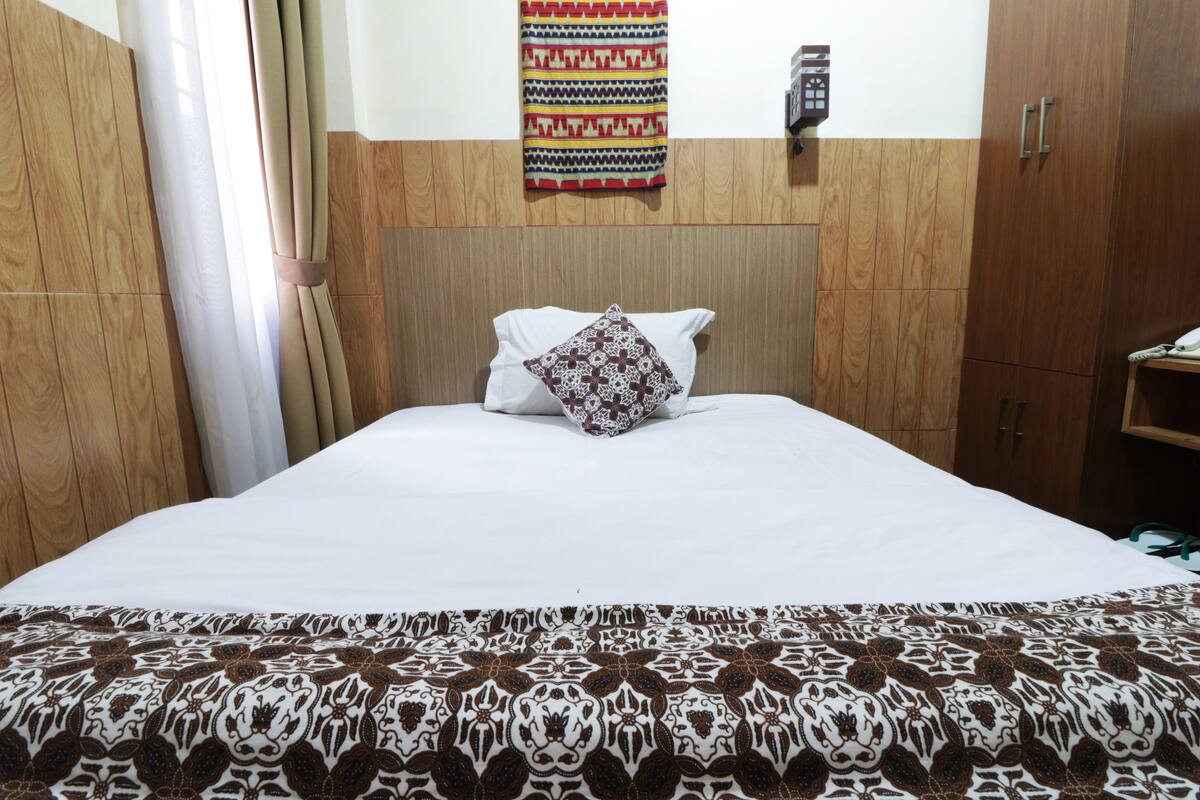 A hotel room with a wooden headboard, a patterned bedspread, a single white pillow, and a wall-mounted telephone.