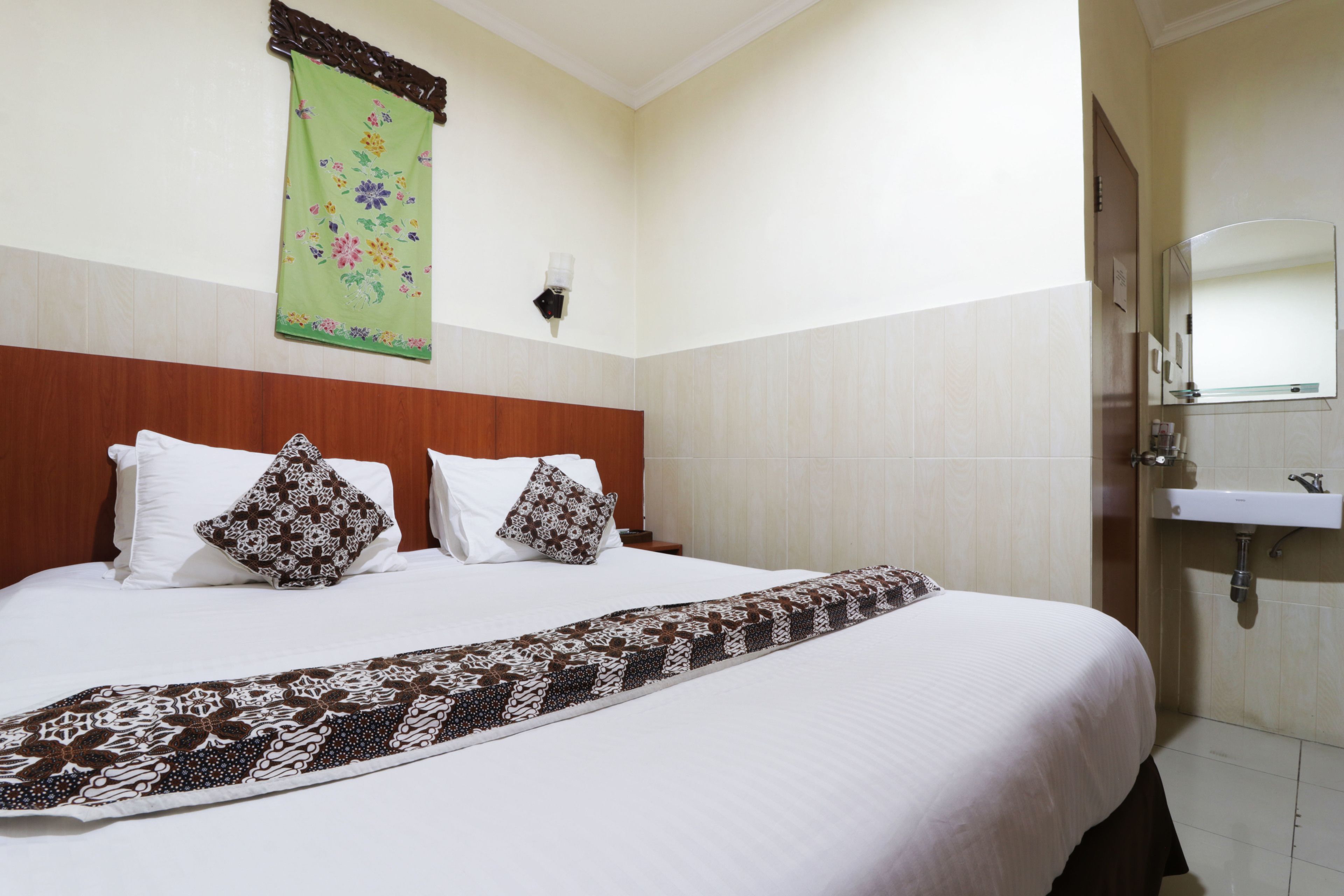 Ein Hotelzimmer mit einem Bett, einer floralen Tapete in Grün, einem hölzernen Kopfende, einem weißen Nachttisch, einem Beistelltisch, einem weißen Waschbecken und einem Spiegel.