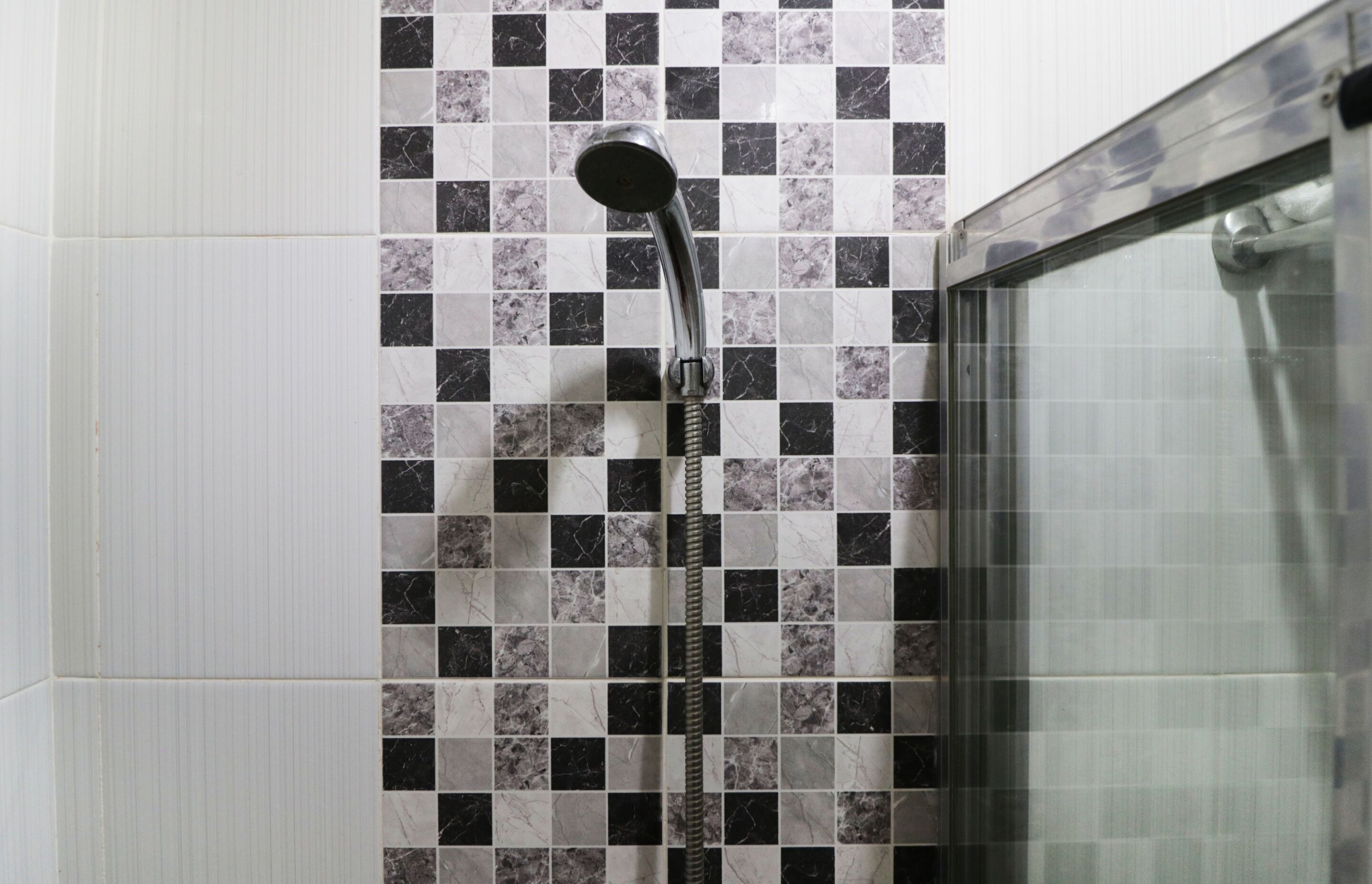 Ducha con cabezal cromado y manguera flexible, frente a una pared de azulejos con un diseño de tablero de ajedrez en blanco y negro.