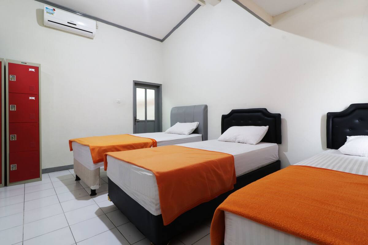 A hotel room with three beds, each with an orange blanket, white bedding, and a grey headboard. There is a red wardrobe and a window with a closed blind.