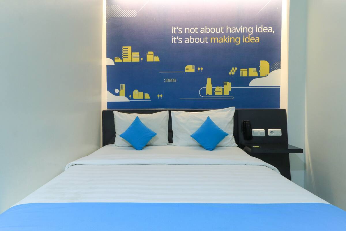 A hotel room with a bed, two blue pillows, a nightstand with a phone, and a wall with a cityscape and motivational quote.
