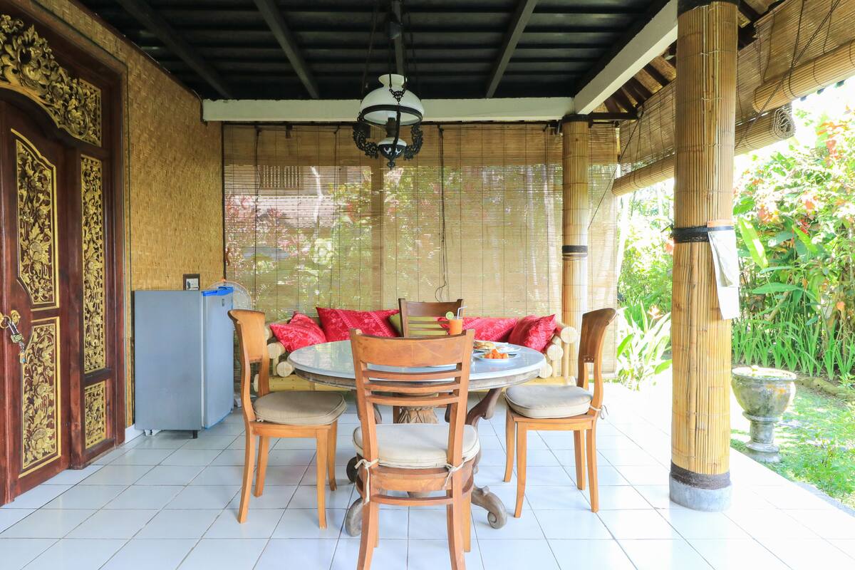 Un espacio comedor al aire libre techado, con una mesa redonda, sillas y un refrigerador.