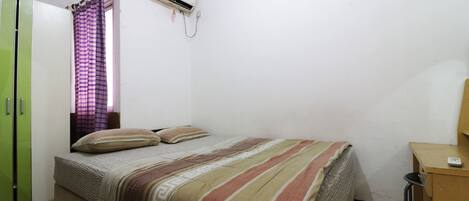 A small, clean room with a bed, a desk, a green wardrobe, and an air conditioning unit.