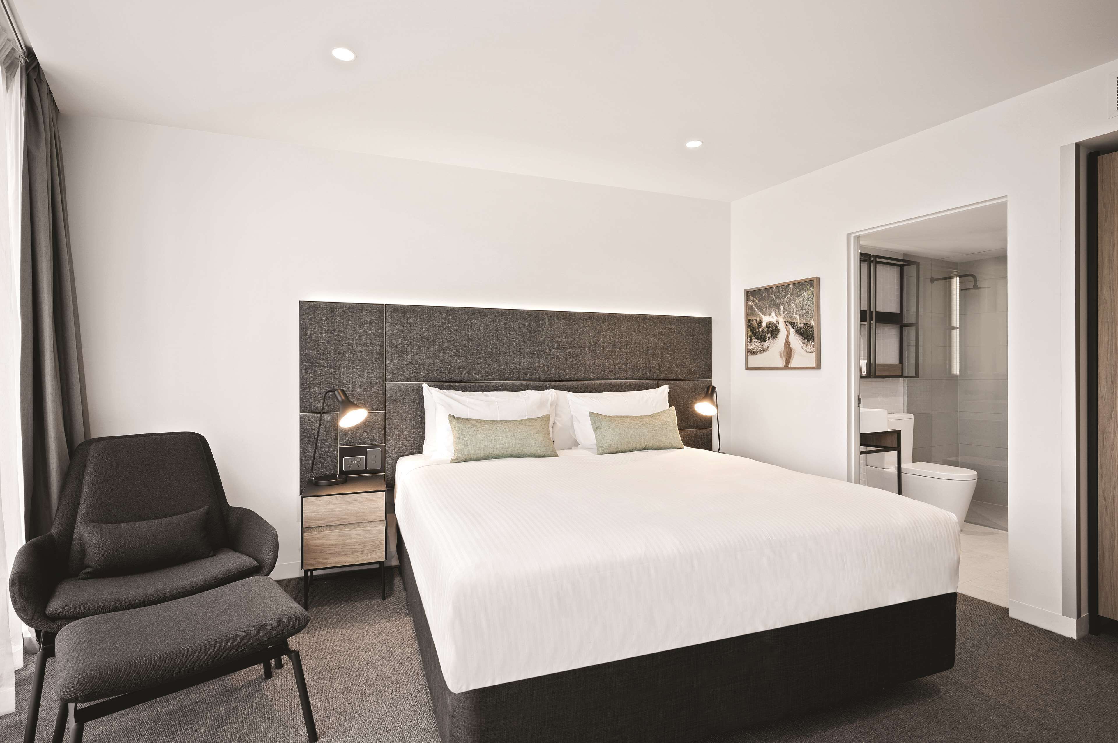 Una habitación de hotel moderna con una cama grande, una silla, un escritorio y un baño visible a través de una puerta abierta.