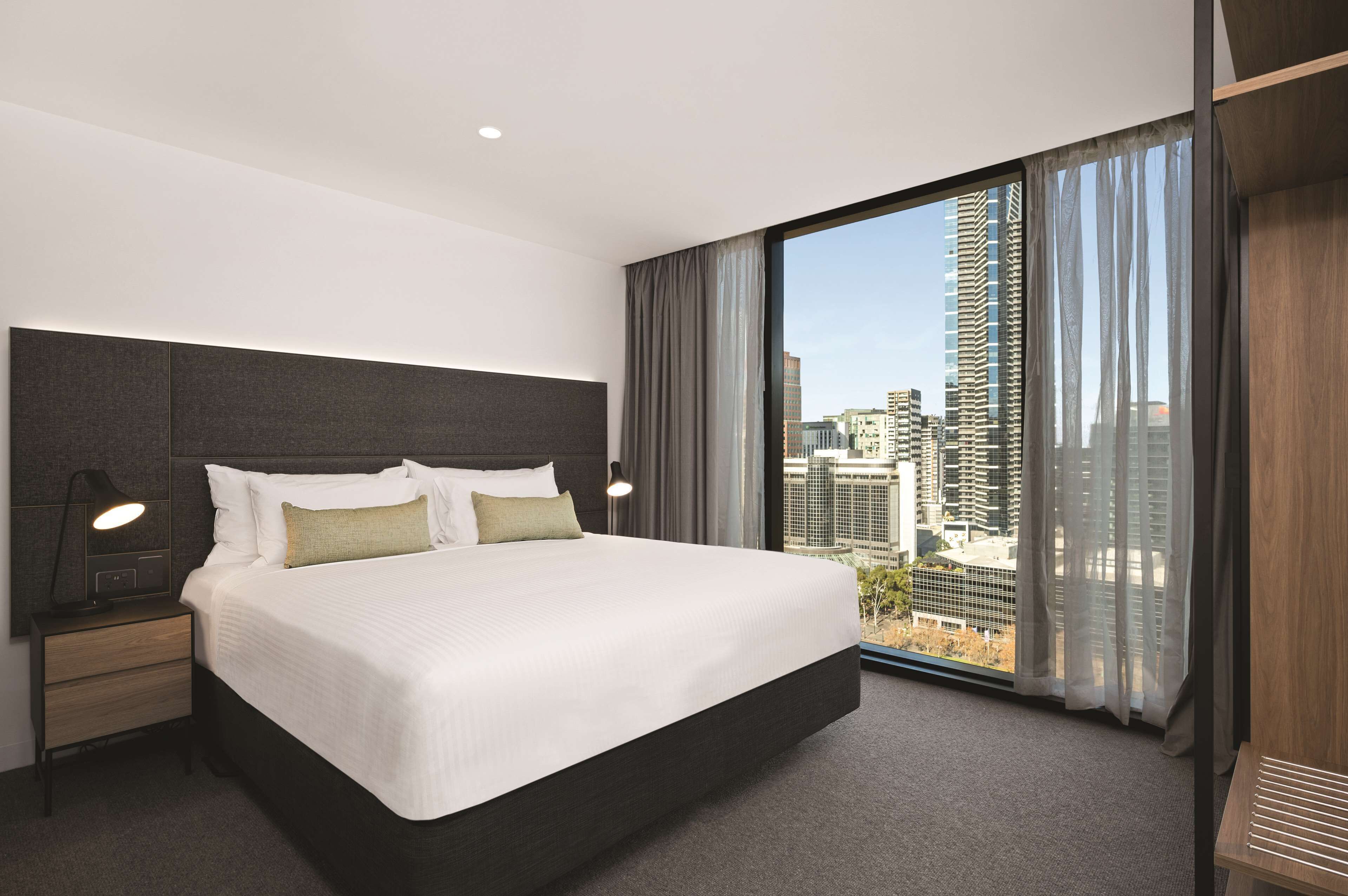 Una habitación de hotel moderna con una cama grande, una mesita de noche, una lámpara de escritorio y vistas de la ciudad a través de un ventanal amplio.