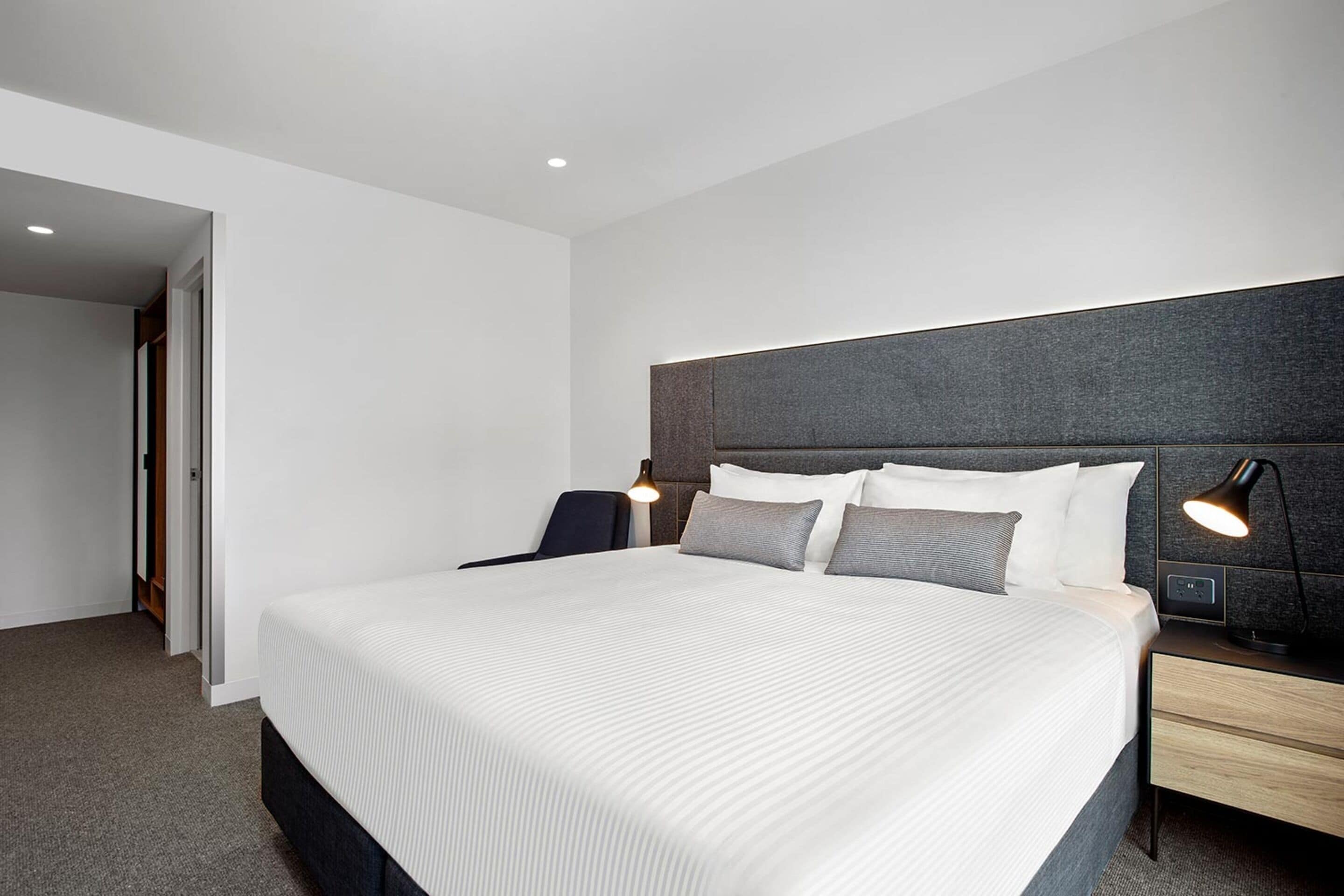 A modern hotel room with a large bed, a desk, and a chair. There is a nightstand with a lamp and a power outlet.