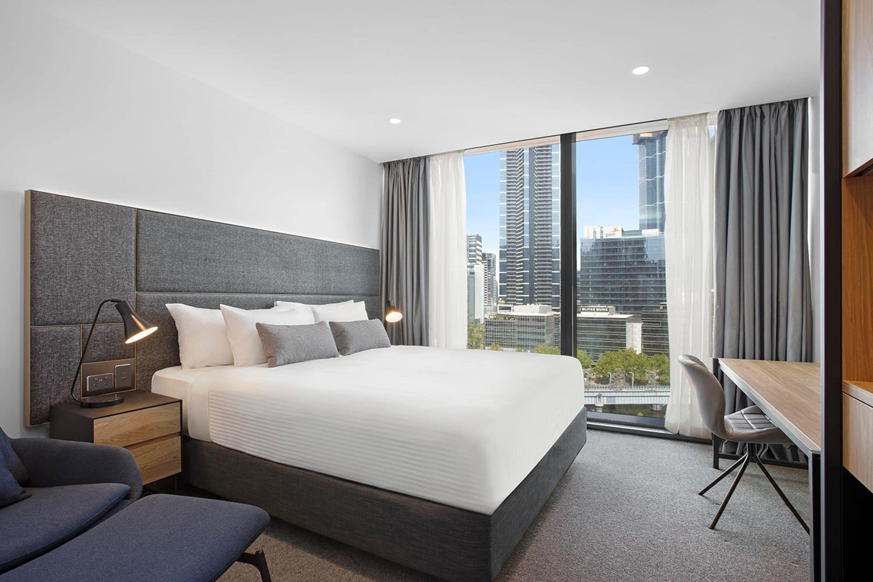 A modern hotel room with a large bed, a desk, and a view of the cityscape.