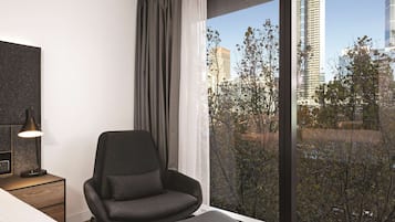 A modern hotel room with a large window, a black chair, a bedside table with a lamp, and a view of city buildings and trees.