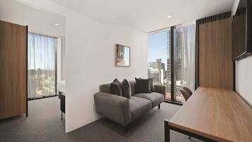 A modern living room with a grey sofa, a wooden coffee table, and a large window offering a city view.