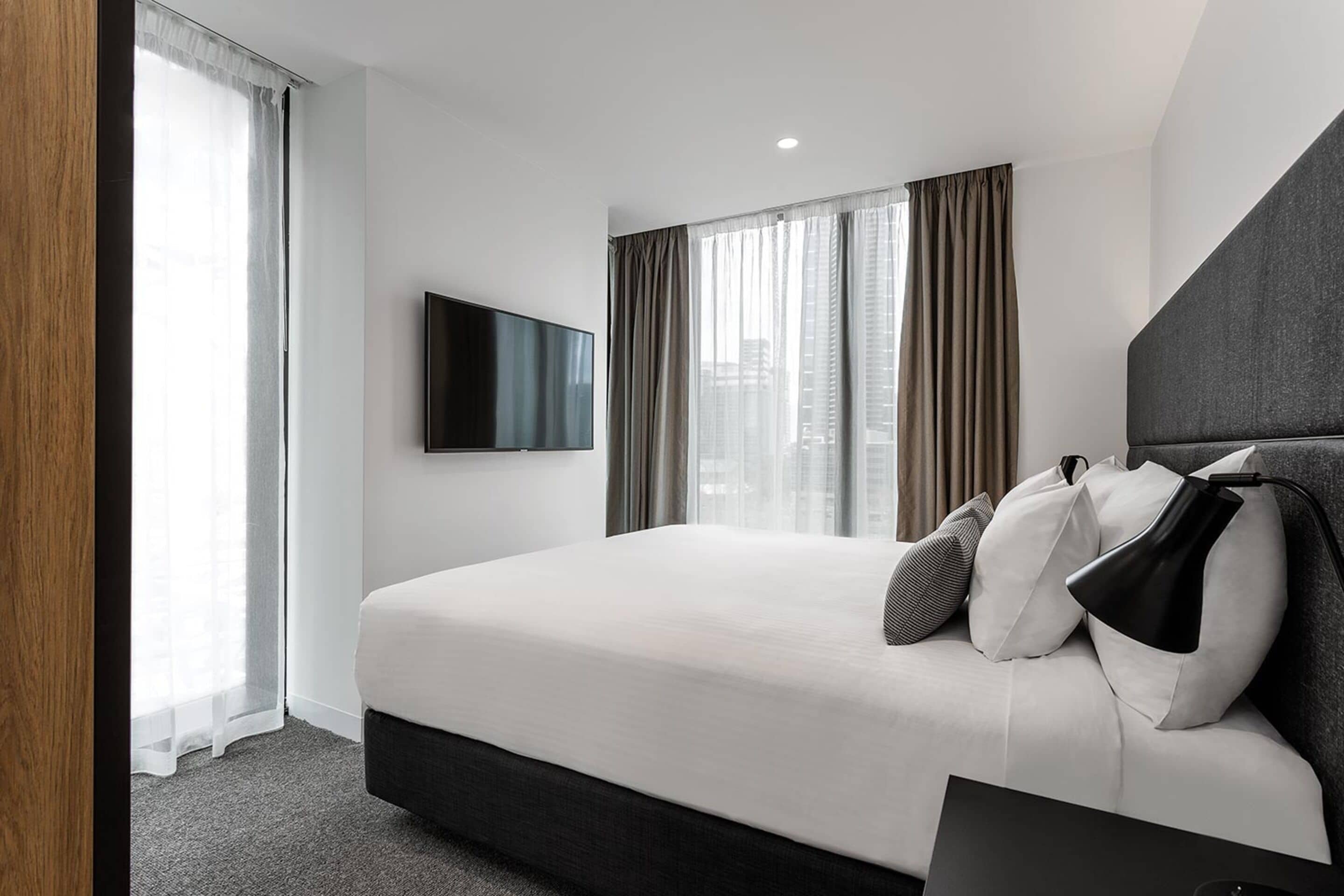 A hotel room with a large bed, a flat-screen TV mounted on the wall, and a view of the city through the window.