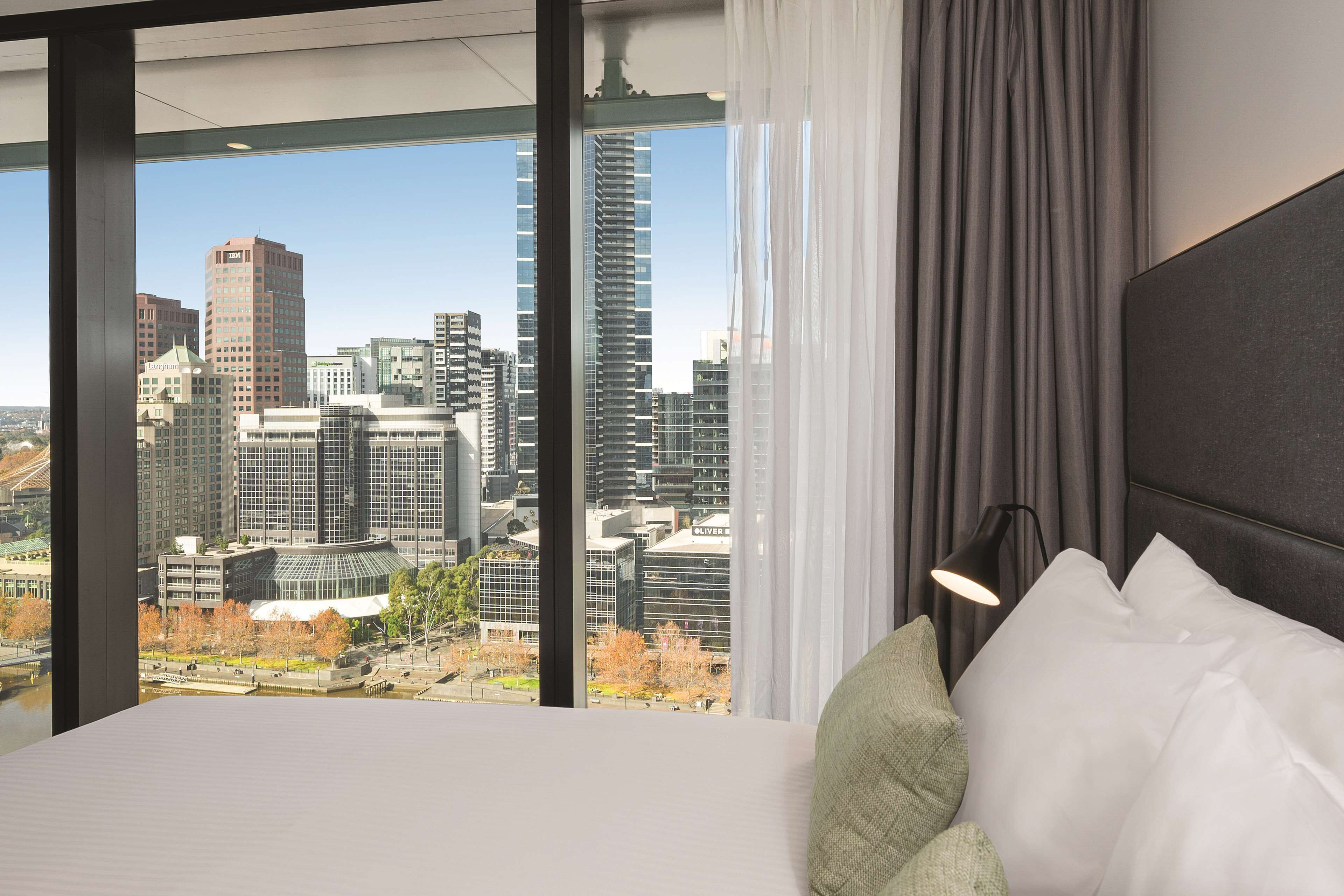 Quarto de hotel com uma janela grande que oferece vista para a cidade, cama com roupa branca, cabeceira preta e abajur.