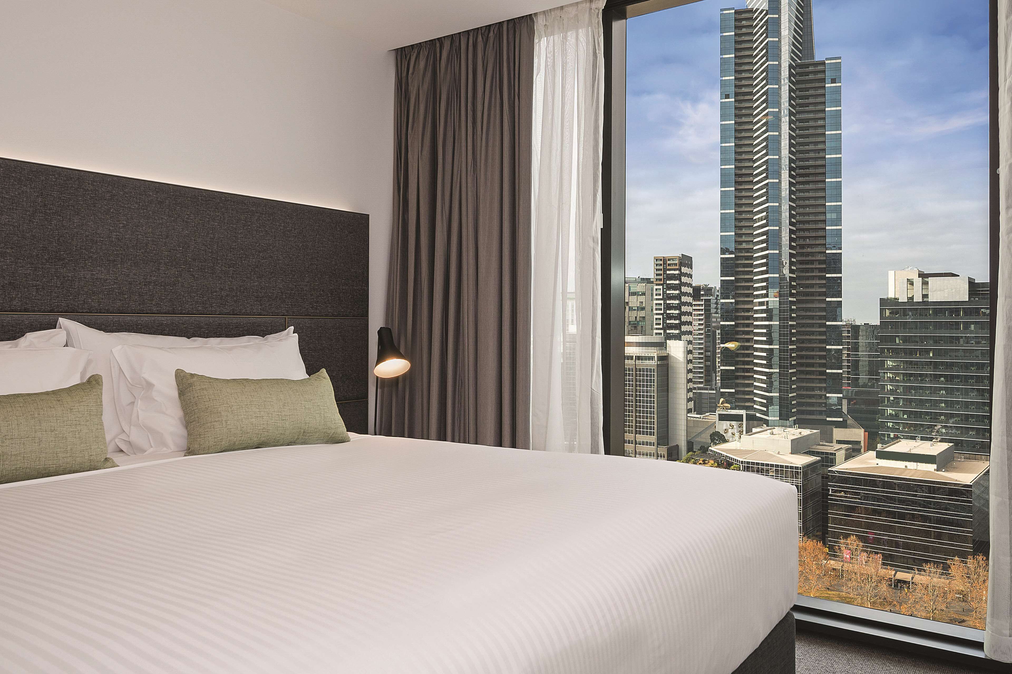 Una habitación de hotel con una cama grande, cabecero, ventana con cortinas y vista a un paisaje urbano.