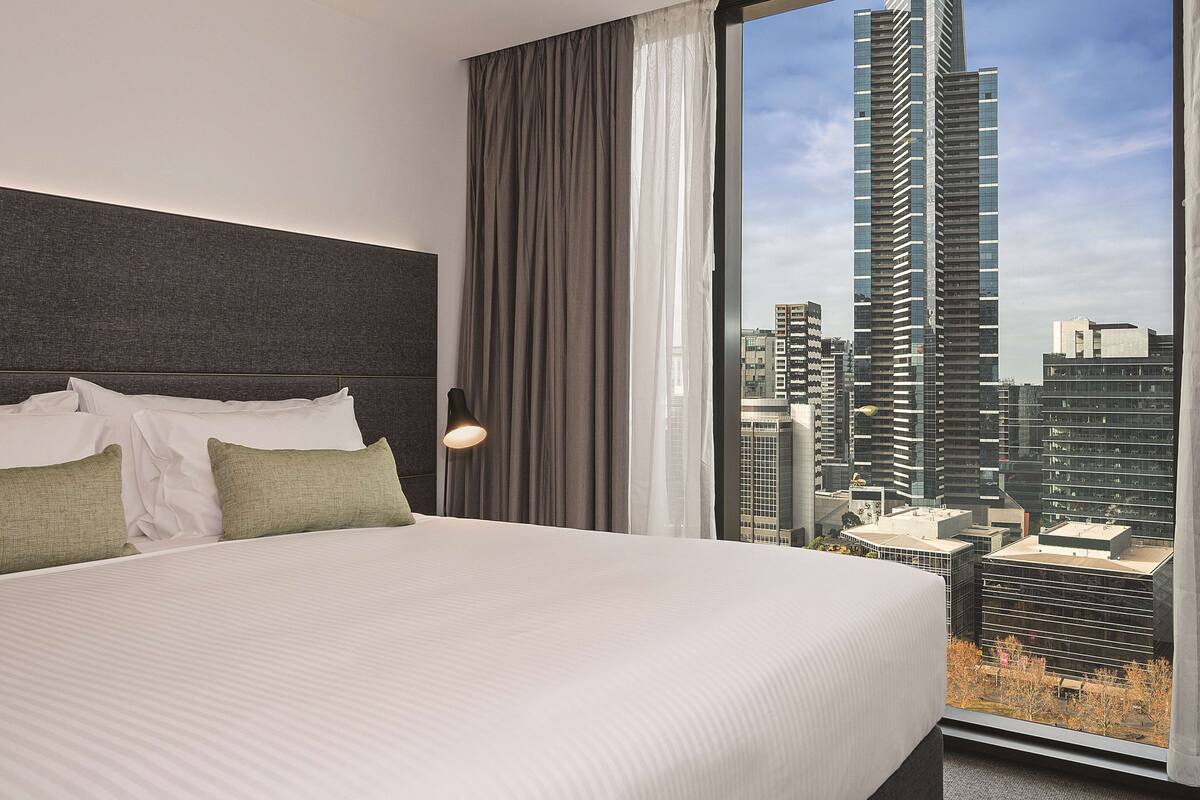 Ein Hotelzimmer mit einem großen Bett, einem Kopfteil, einem Fenster mit Vorhängen und Blick auf eine Stadtlandschaft.