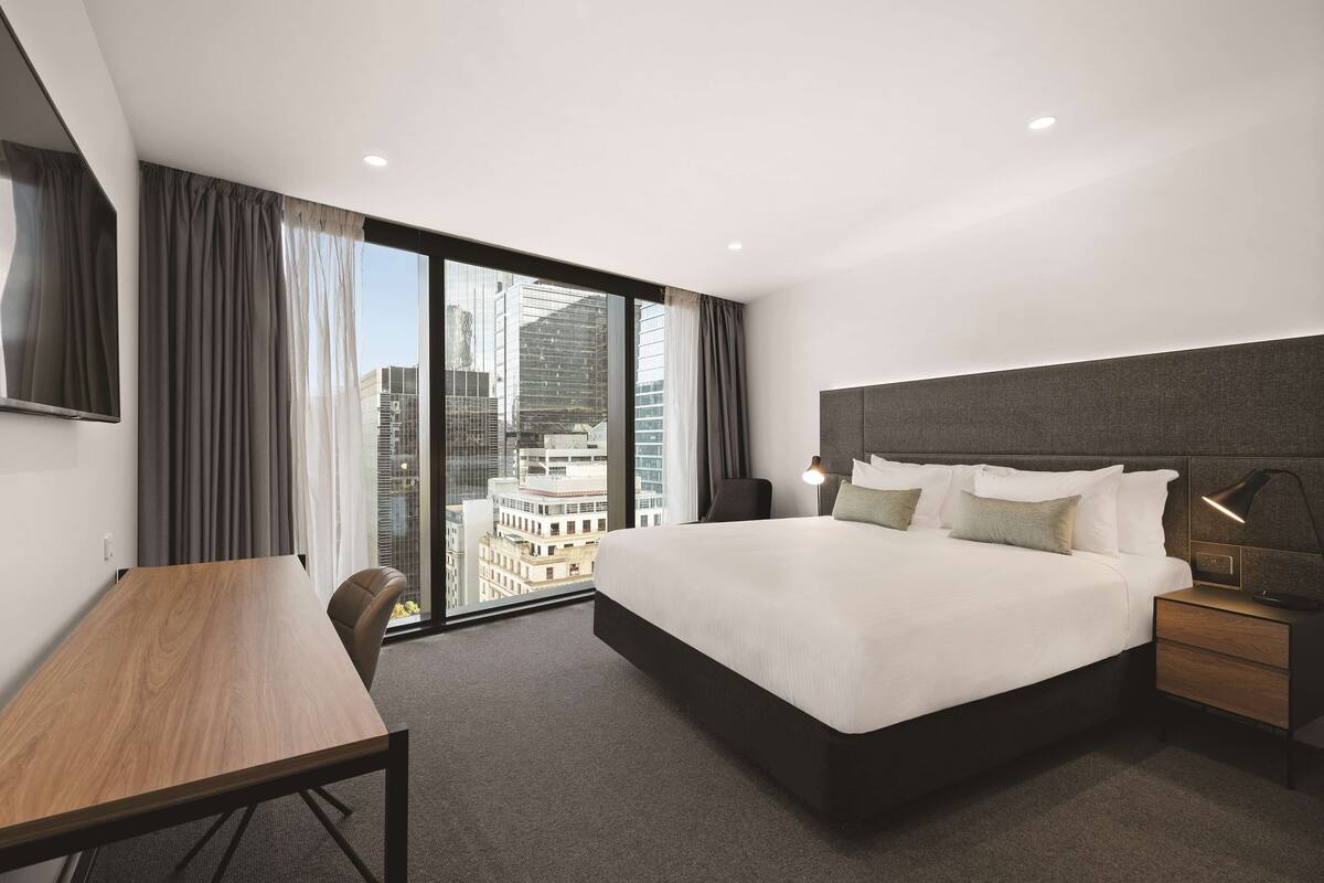 Ein modernes Hotelzimmer mit einem großen Bett, einem Schreibtisch und Blick auf die Stadt durch raumhohe Fenster.