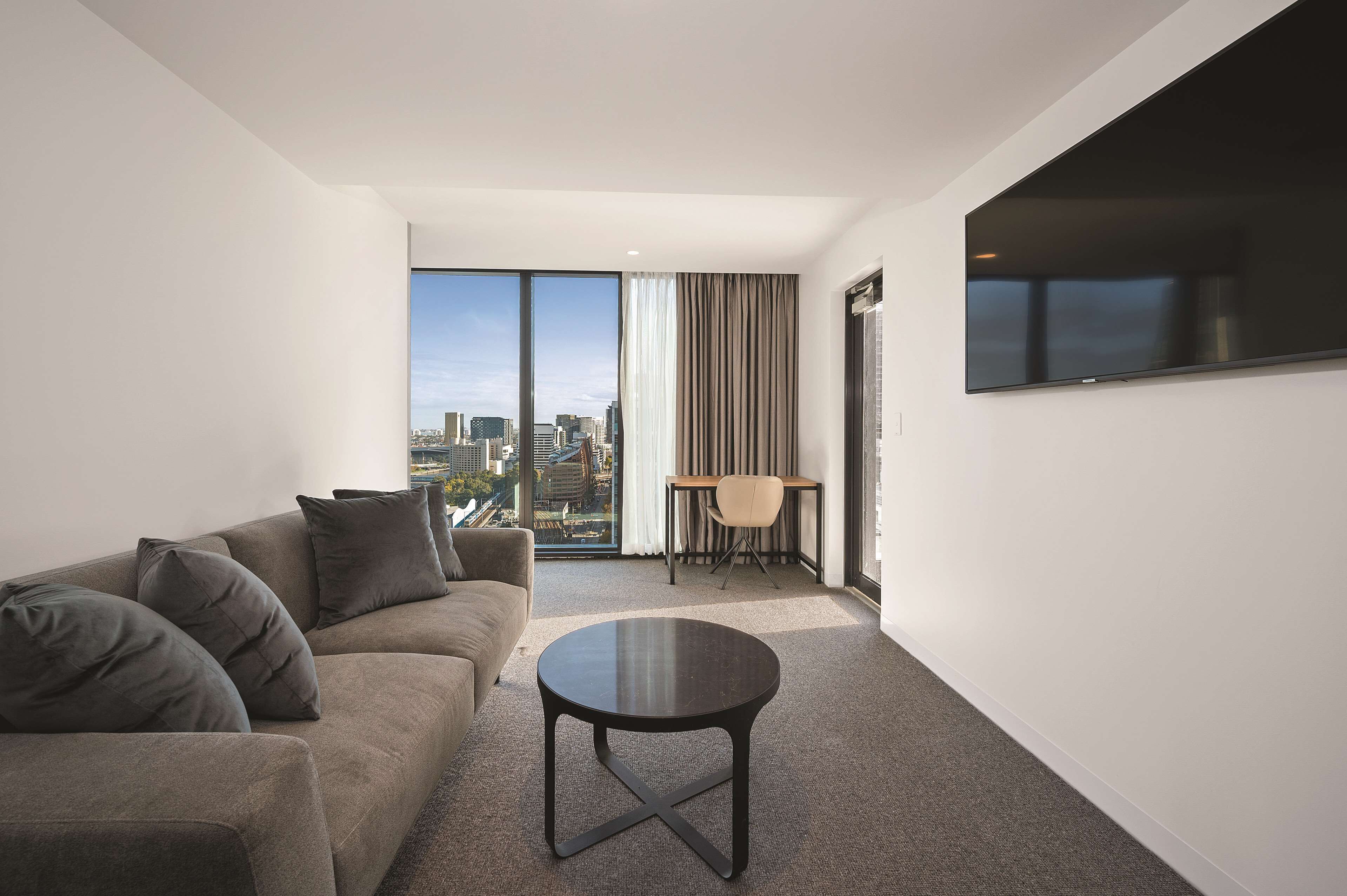 A modern hotel room with a large window, a sofa, a round table, and a TV mounted on the wall.