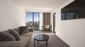 A modern hotel room with a large window, a sofa, a round table, and a TV mounted on the wall.