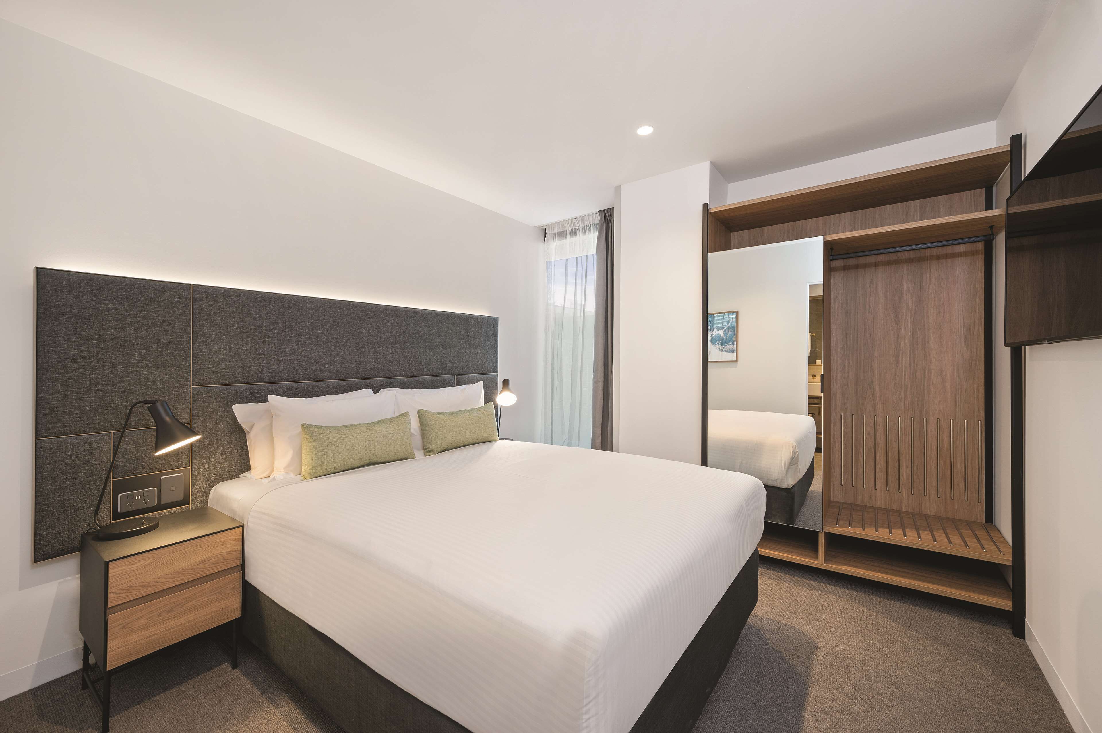 Una habitación de hotel moderna con una cama grande, una mesita de noche con lámpara, un armario y un espejo.