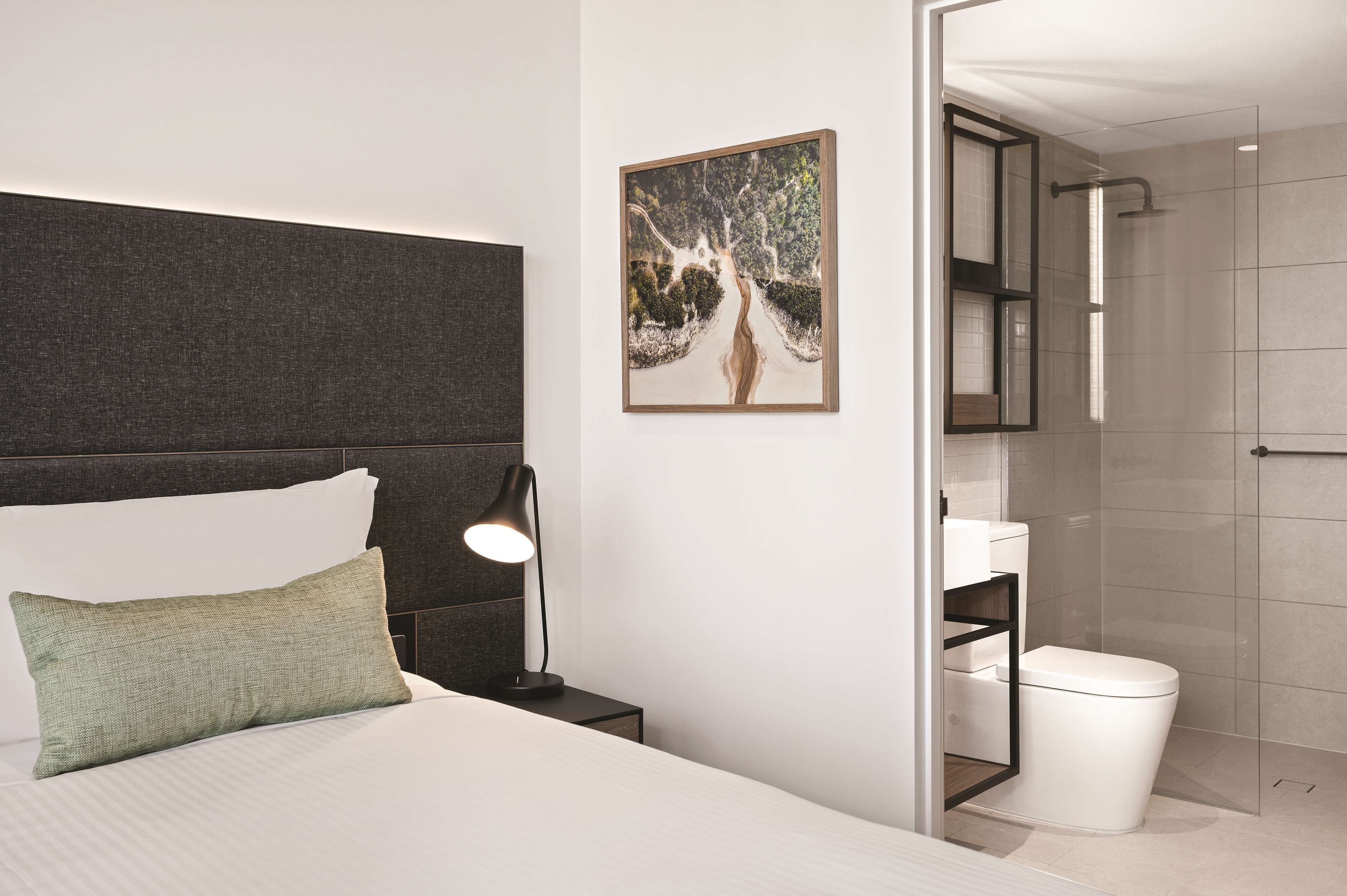 Una habitación de hotel moderna con una cama, una mesita de noche, una lámpara de noche, un cuadro enmarcado y una zona de ducha.