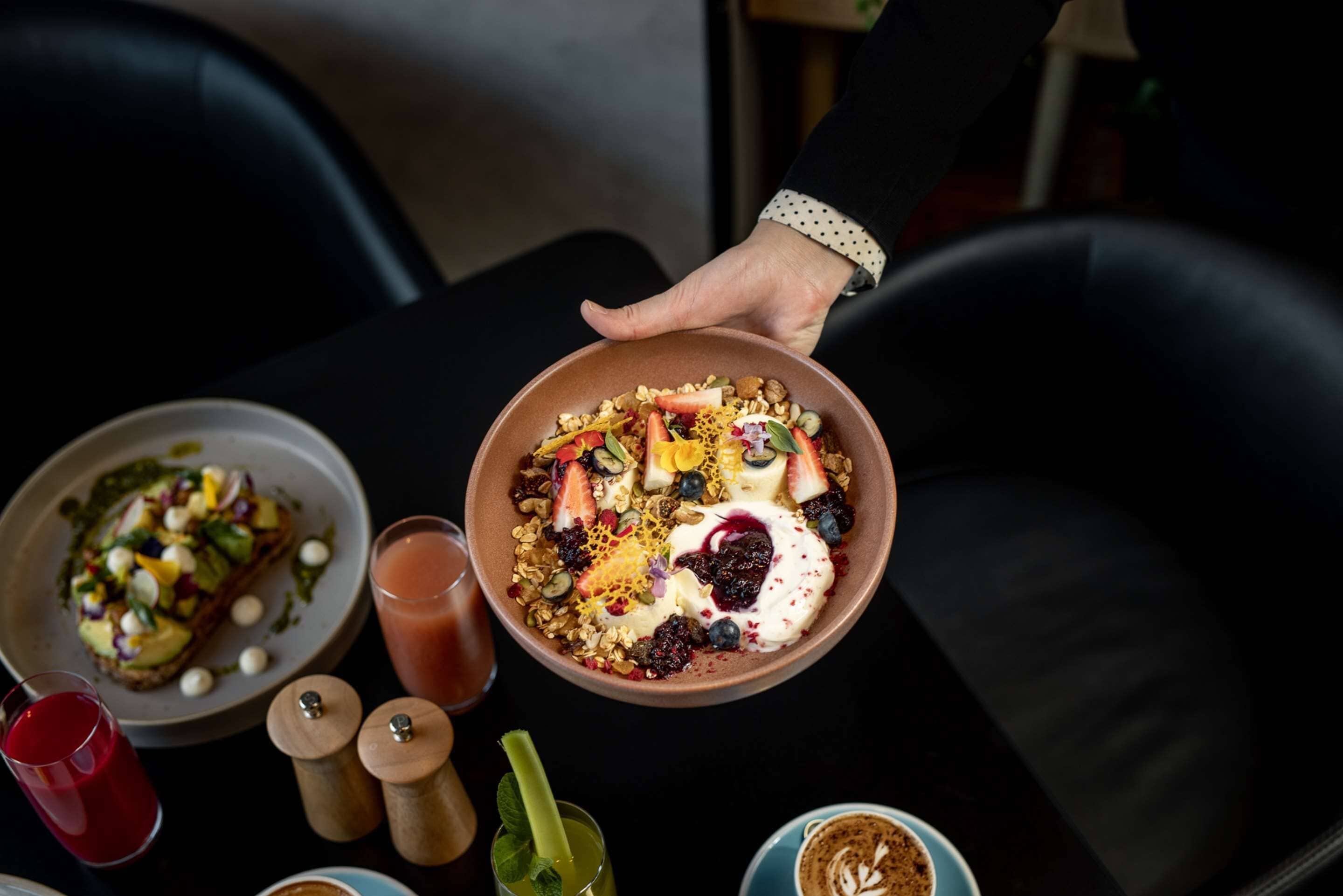 En person holder en skål med morgenmad bestående af forskellig frugt og granola, et glas rød juice og en kop kaffe på et bord.
