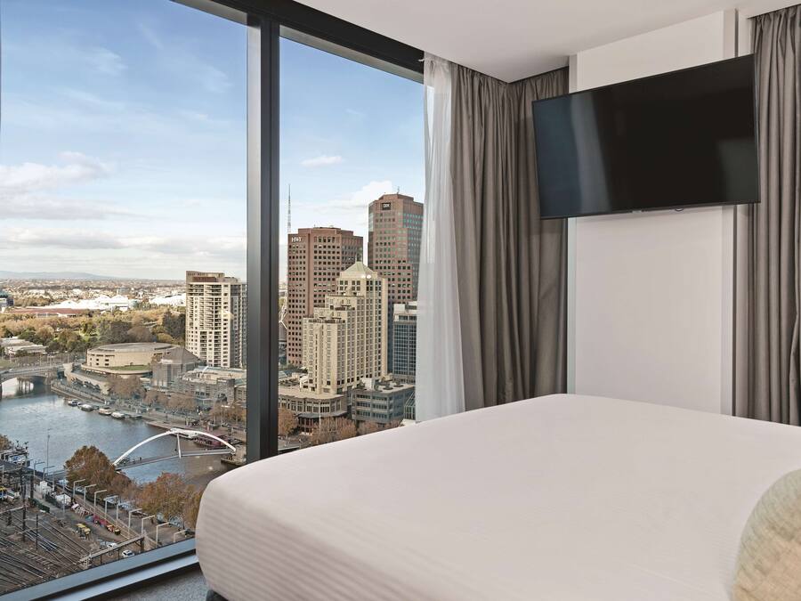 A hotel room with a large bed, a flat-screen TV, and a view of a cityscape including a river and bridges.