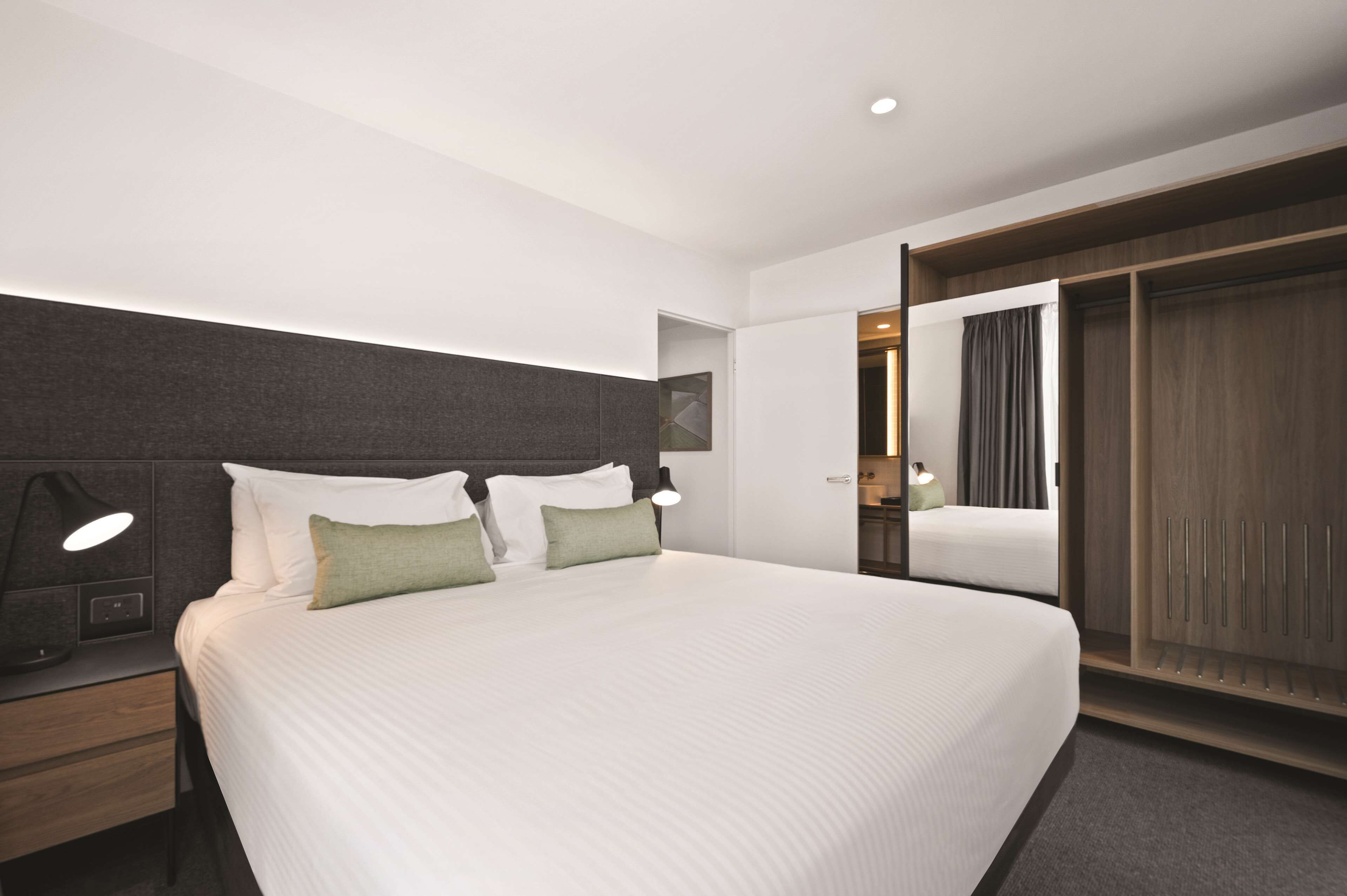 Una habitación de hotel moderna con una cama grande, una mesita de noche con lámpara, un armario y un espejo.