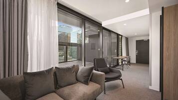 A modern hotel room with a sofa, chairs, a dining table, and large windows offering a city view.