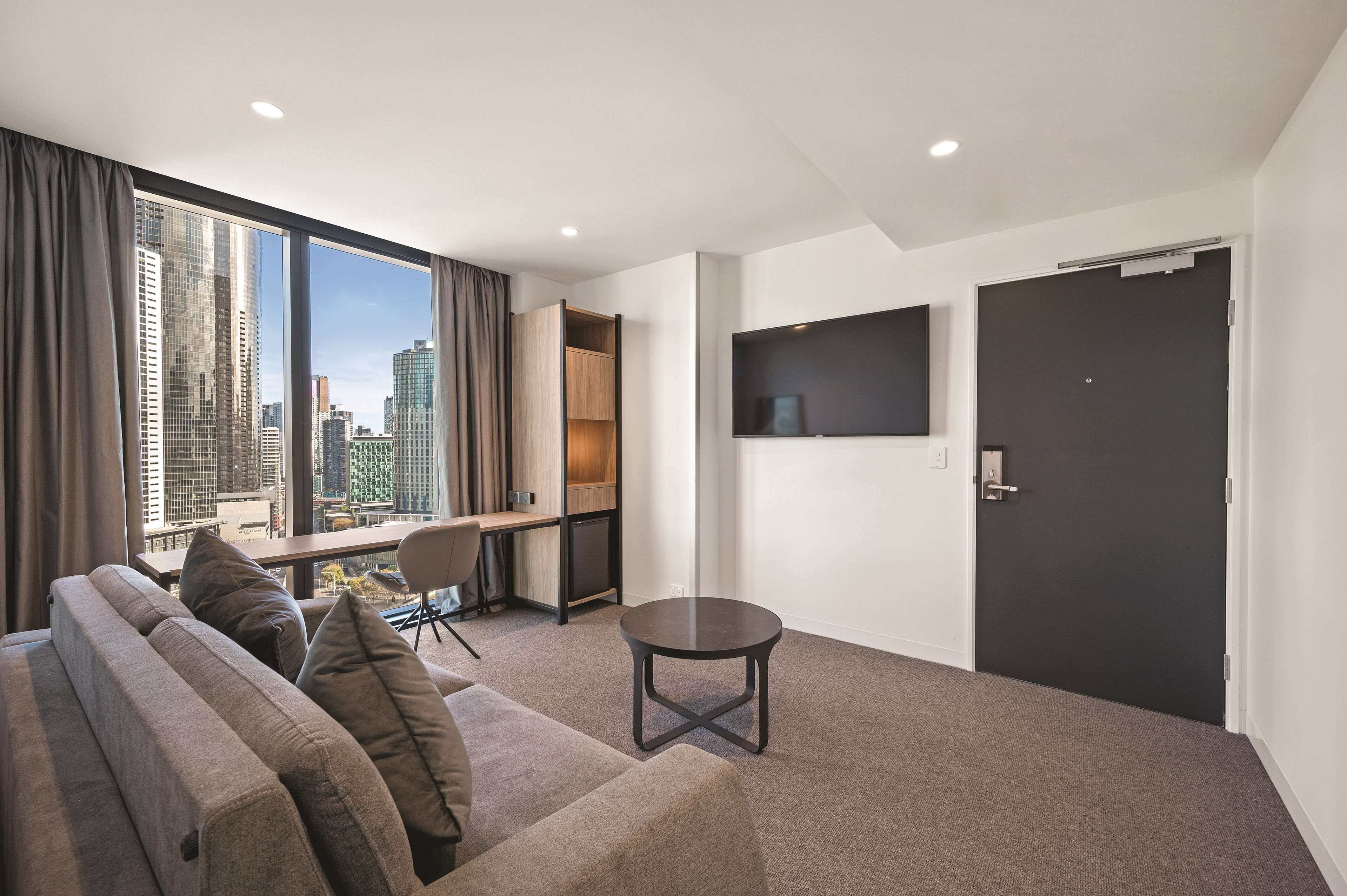 A modern hotel room with a large window offering a city view, a sofa, a small table, and a flat-screen TV mounted on the wall.
