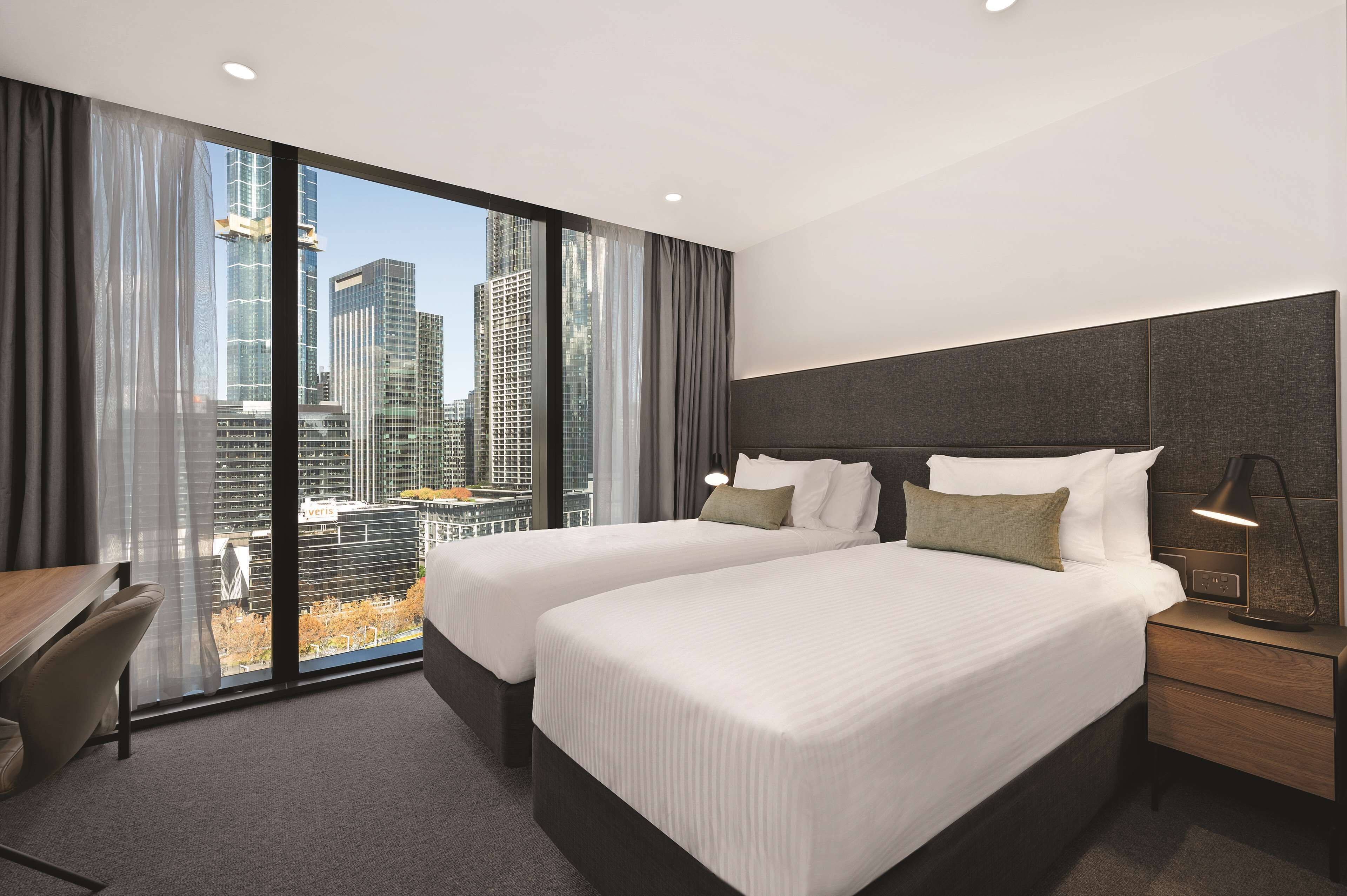 Habitación de hotel con dos camas, un escritorio y vistas a edificios de la ciudad.