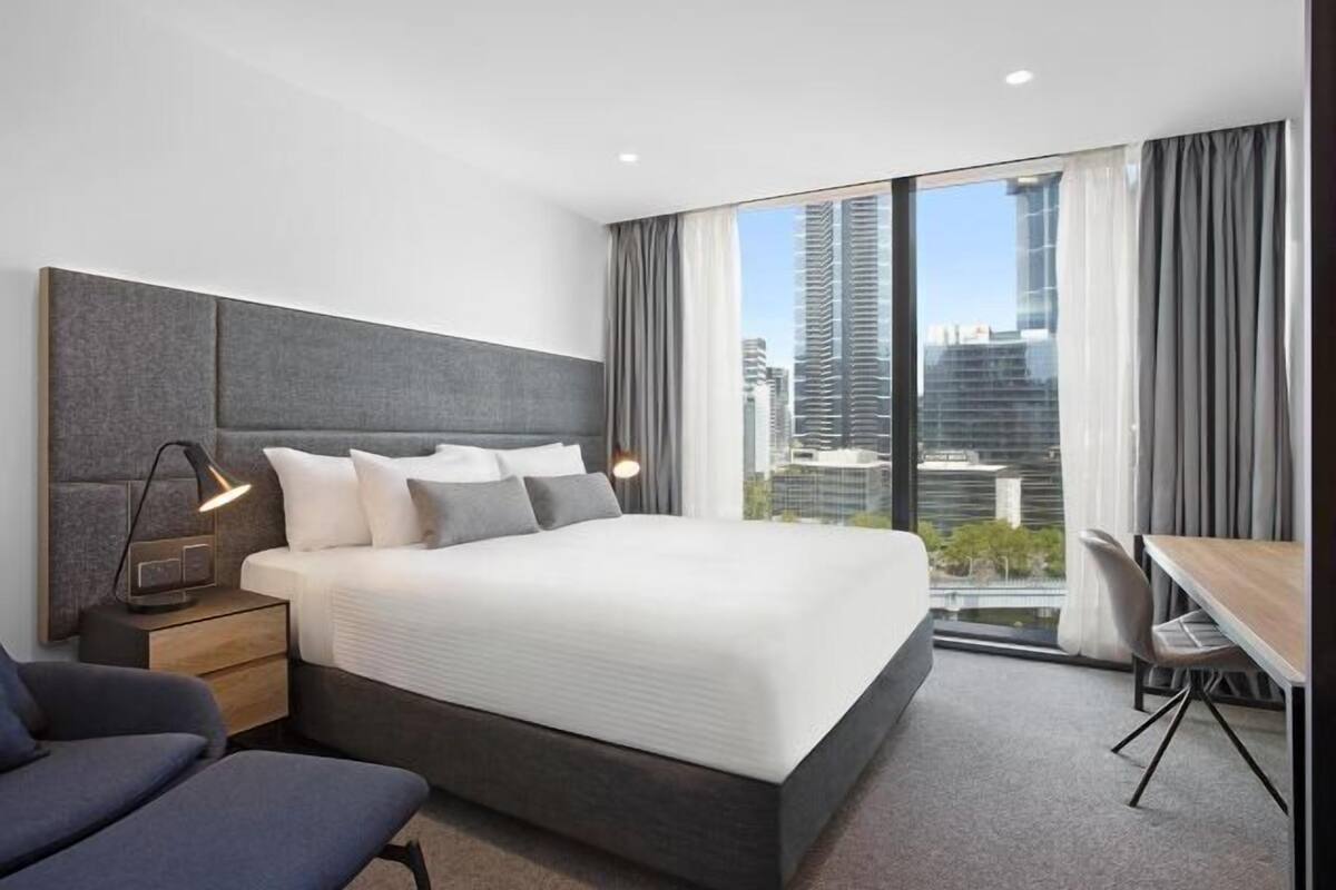 Ein modernes Hotelzimmer mit einem großen Bett, Nachttischen, einem Schreibtisch und Blick auf die Stadtlandschaft.