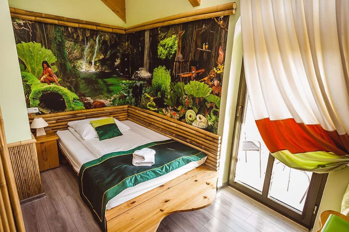 Una habitación con cama, mesita de noche, ventana con cortinas y un mural con paisaje de bosque.