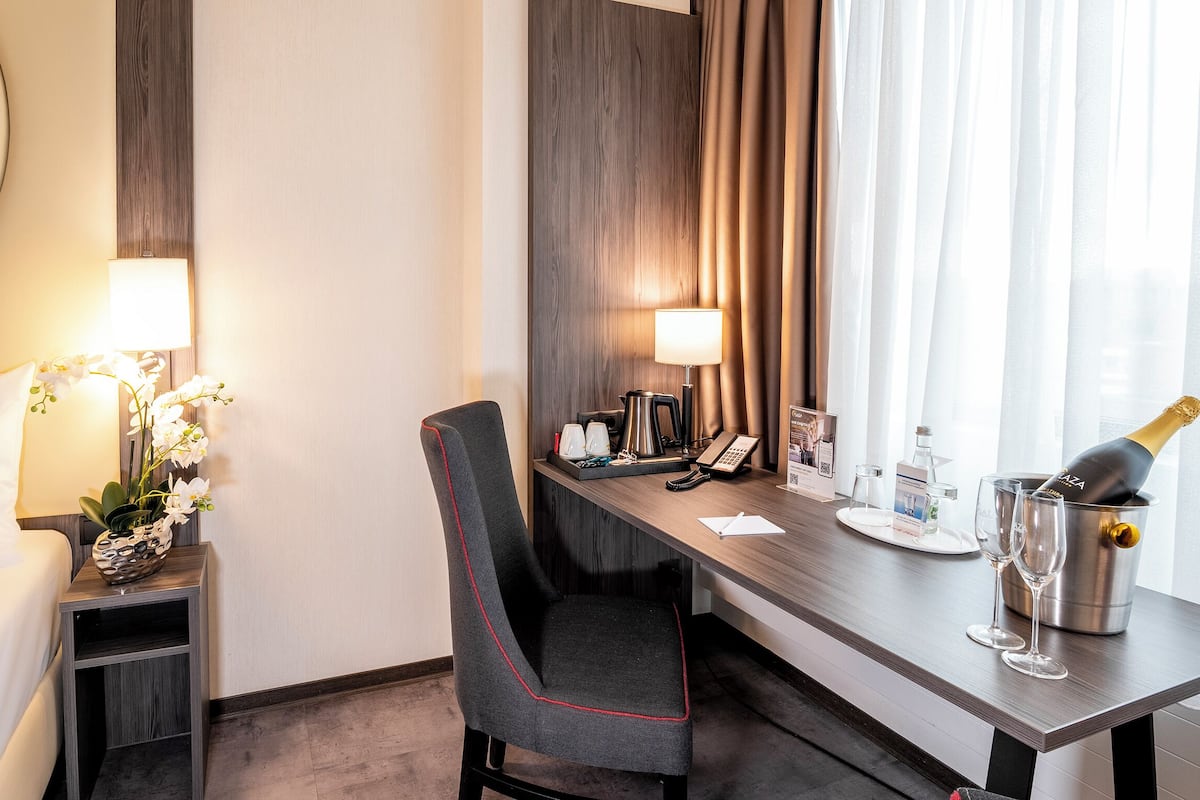 A hotel room with a bed, a desk with a lamp, a chair, a minibar, and a bottle of champagne.