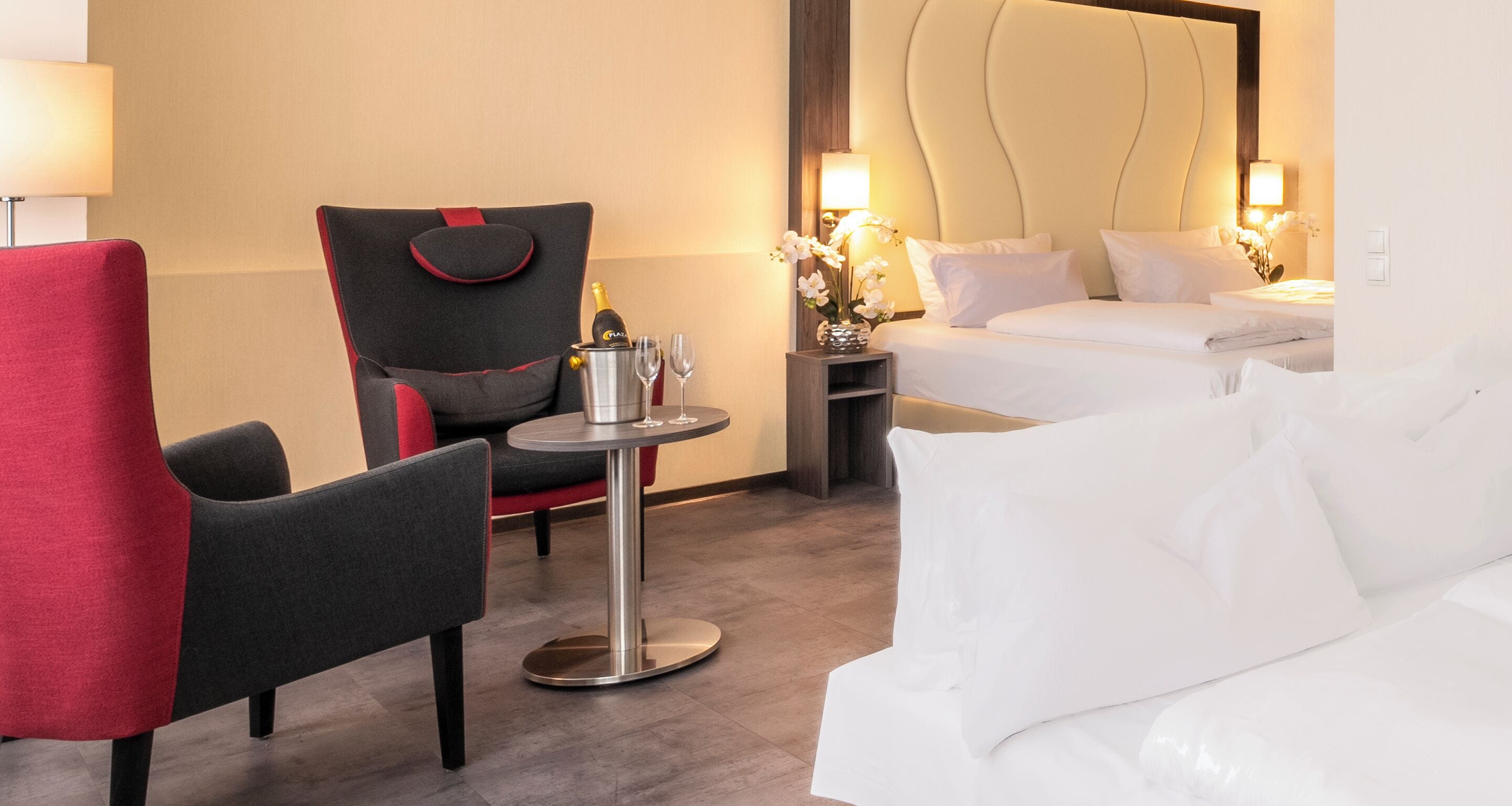 A hotel room with a bed, a red and black chair, a small table with a bottle and glasses, and a nightstand with a lamp.