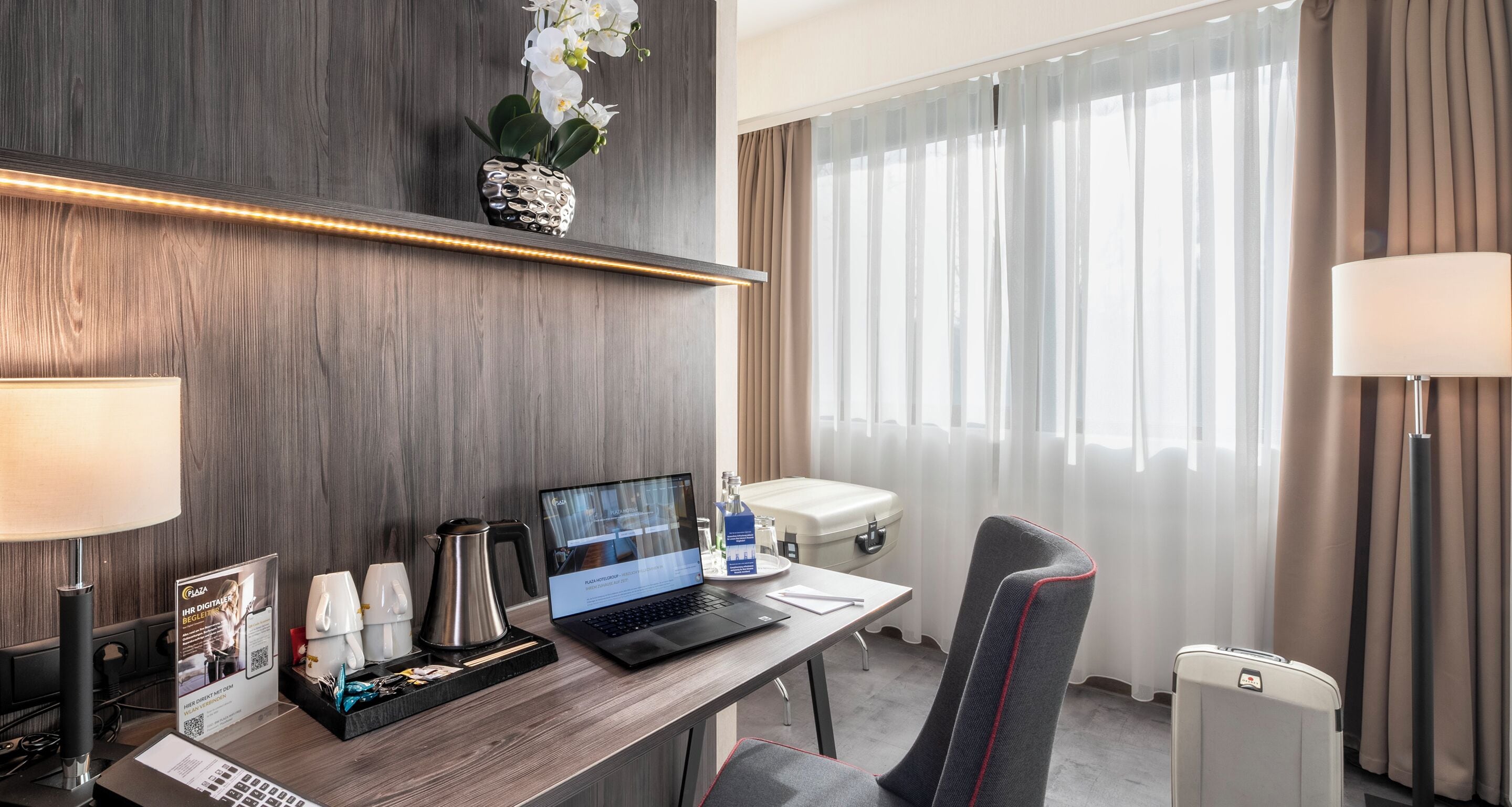 A hotel room with a wooden desk, a laptop, a coffee maker, a chair, a bedside table, a lamp, a window with curtains, and a potted plant.