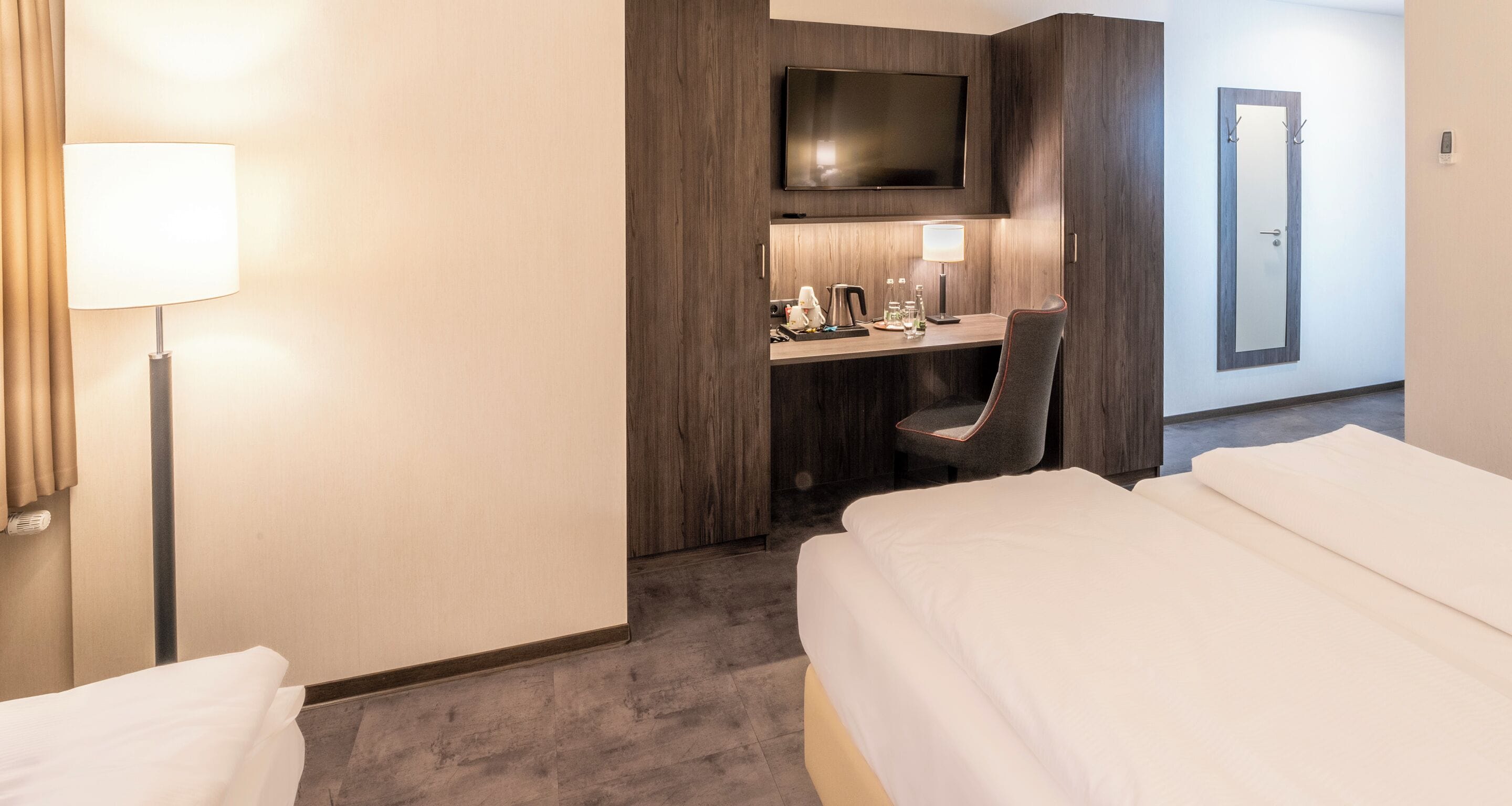 A hotel room with a bed, a desk with a chair, a TV, and a bedside lamp.