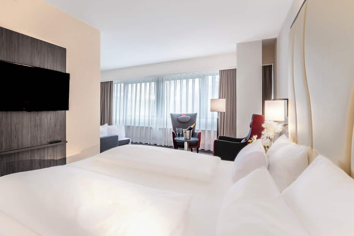 A hotel room with a large bed, a flat-screen TV mounted on the wall, a dining area with a table and chairs, and a view of the city through large windows.