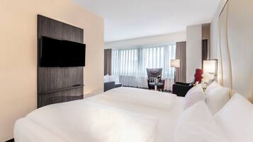 Habitación de hotel con una cama grande, un televisor de pantalla plana montado en la pared, un área de comedor con mesa y sillas, y vistas a la ciudad a través de amplios ventanales.