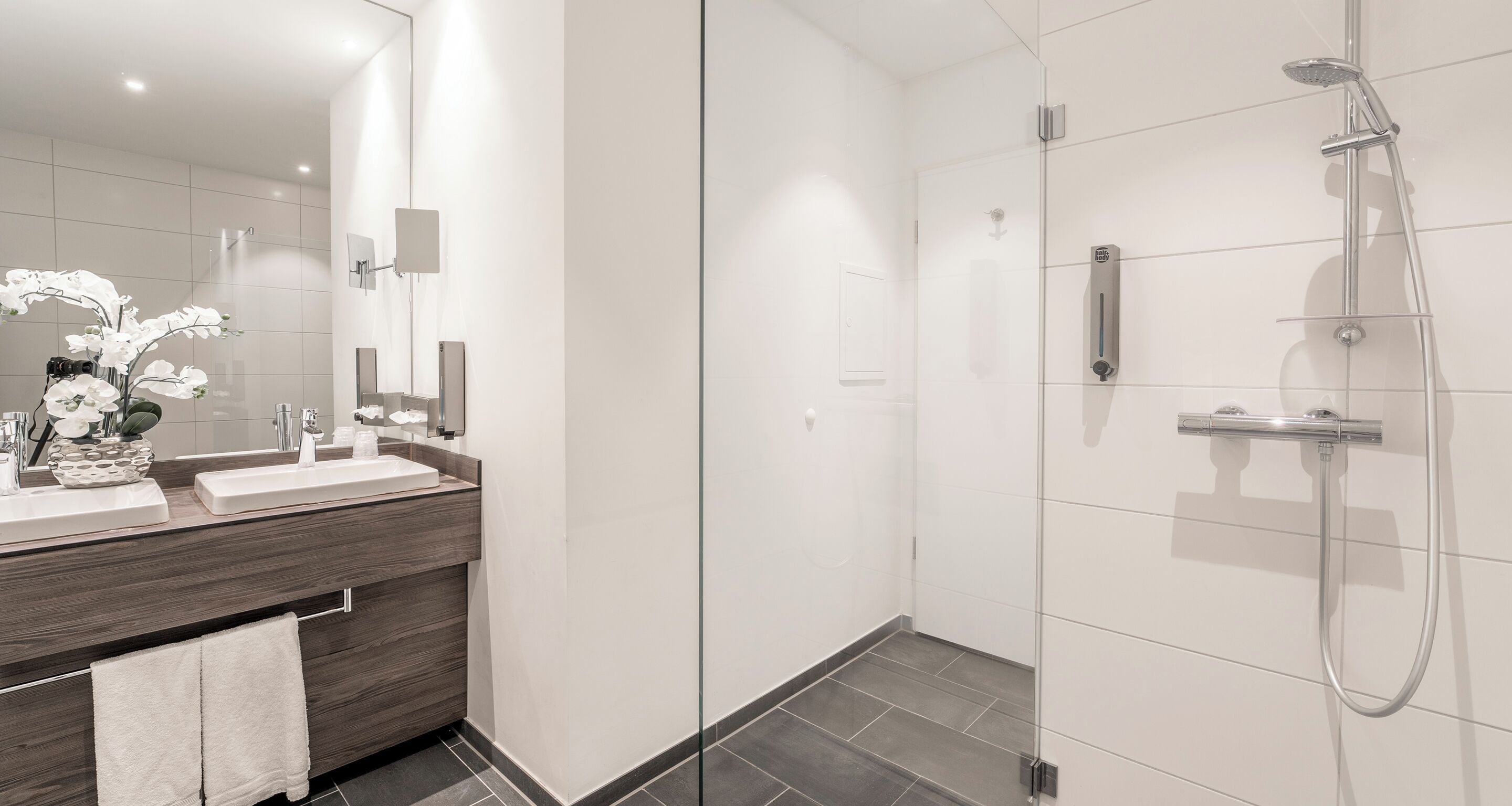 Un baño moderno con cabina de ducha de vidrio, tocador con espejo y un jarrón de flores blancas.